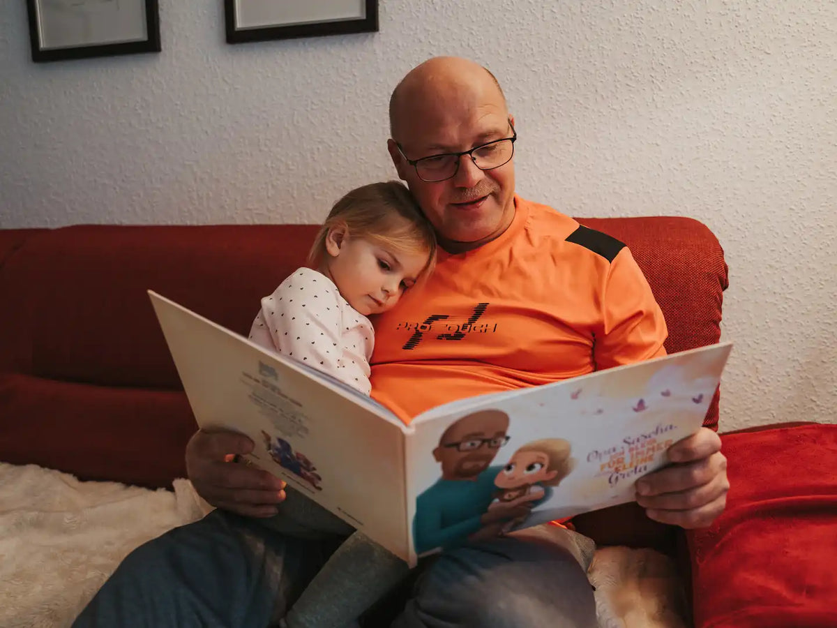 Ein Opa und Enkeltochter kuscheln auf der Couch und lesen im individuellen Buch von Hurra Helden. 
